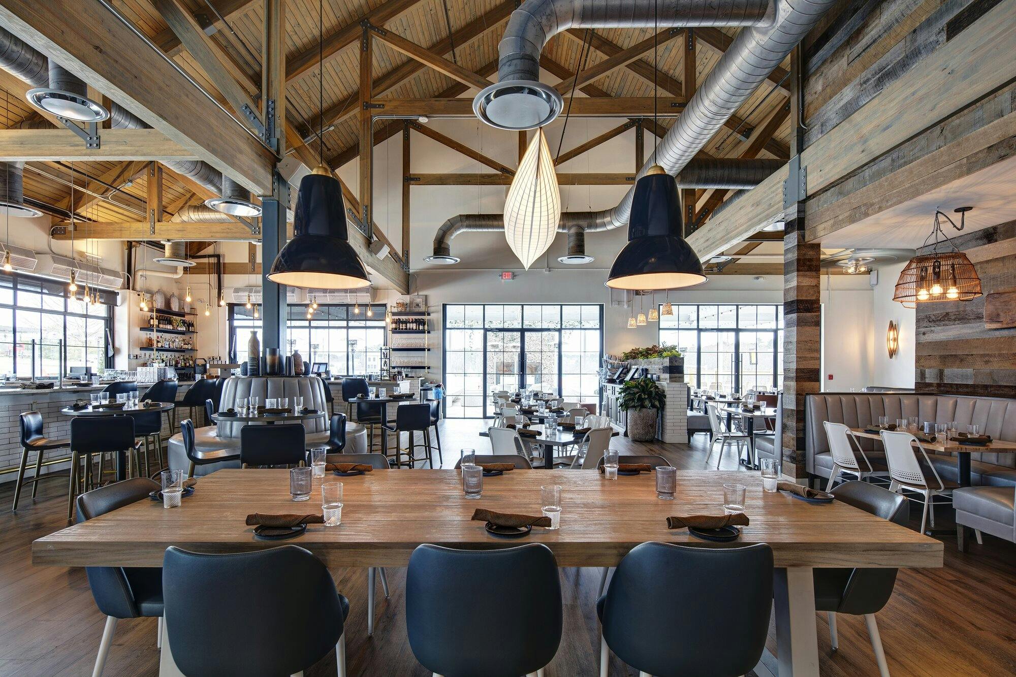 Inside restaurant view with chairs, tables, booths, and bar. Exposed duct work, heavy natural wood beams, and modern hanging light fixtures.