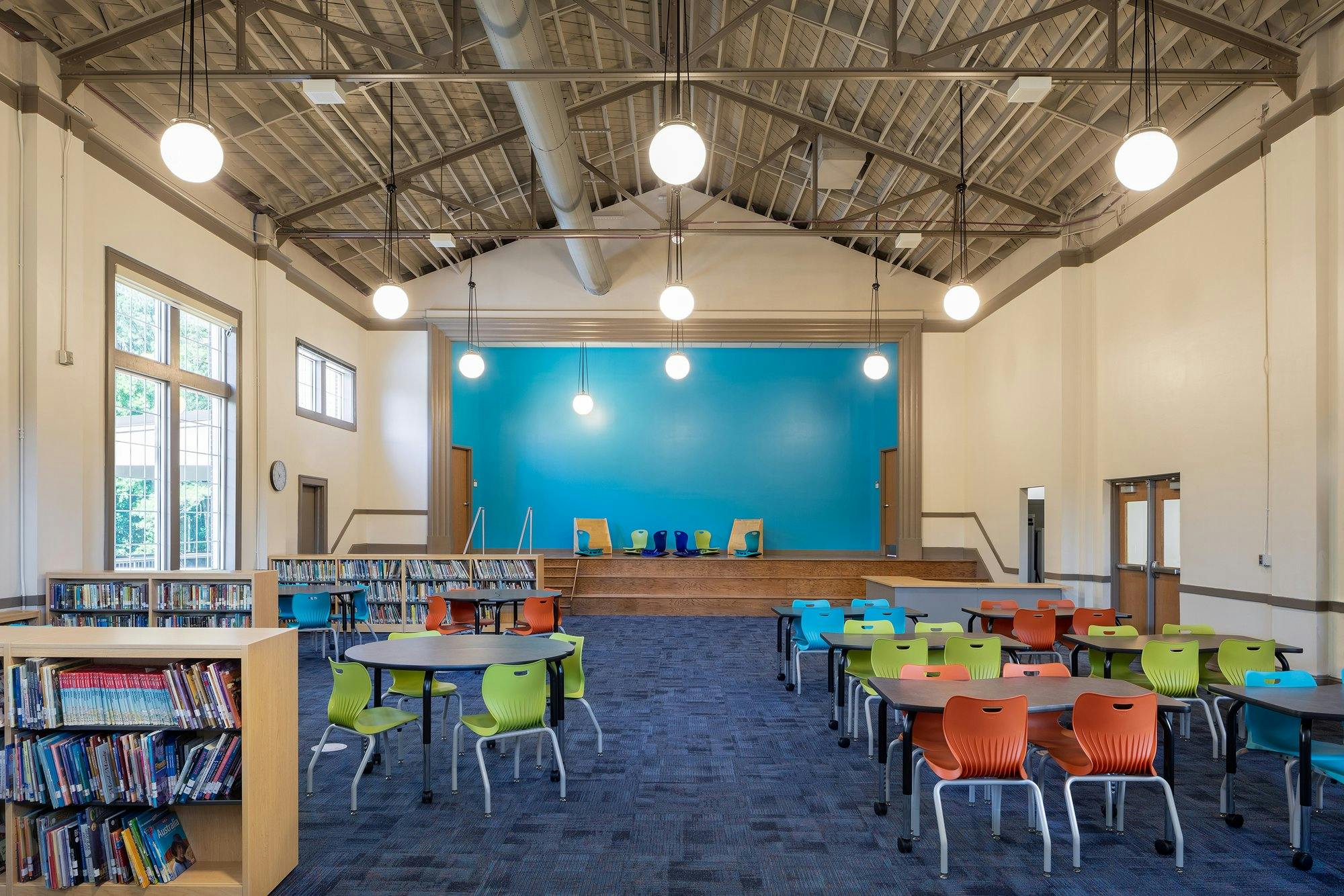 Books can be seen lining short wooden shelves in a library. Blue, green, and orange seats line a blue carpeted floor. 