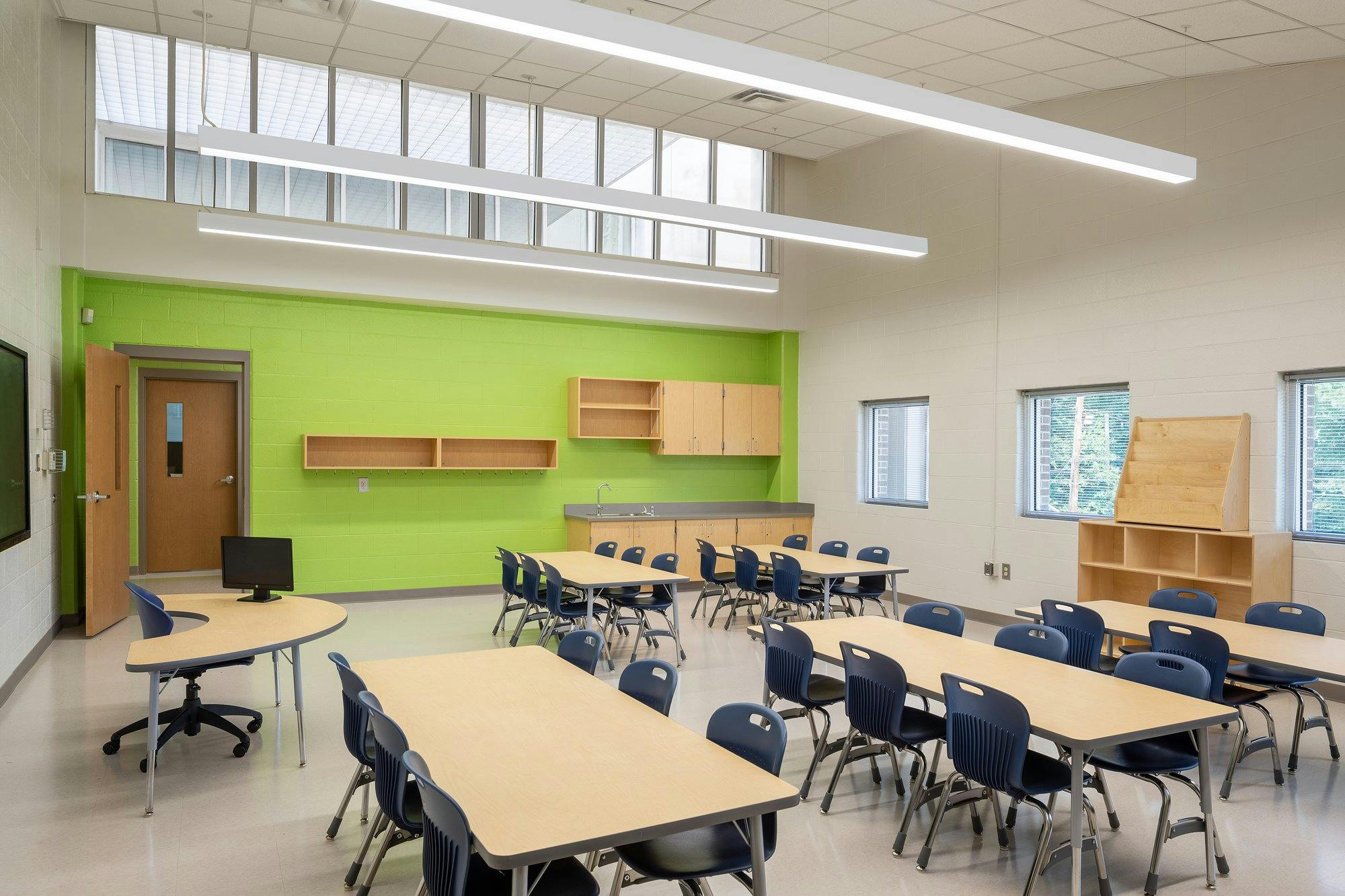 A room with a high ceiling and an electric green accent wall. Tables and chairs fill the floor of the classroom.