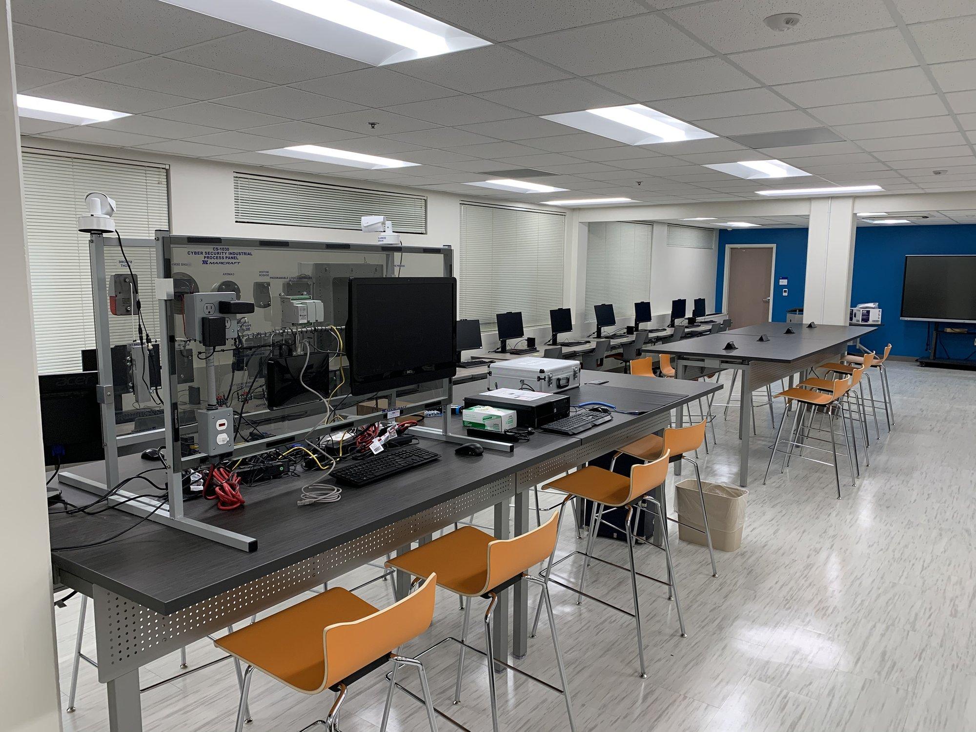 Computer screens and wires fill table tops in a large cybersecurity computer lab.