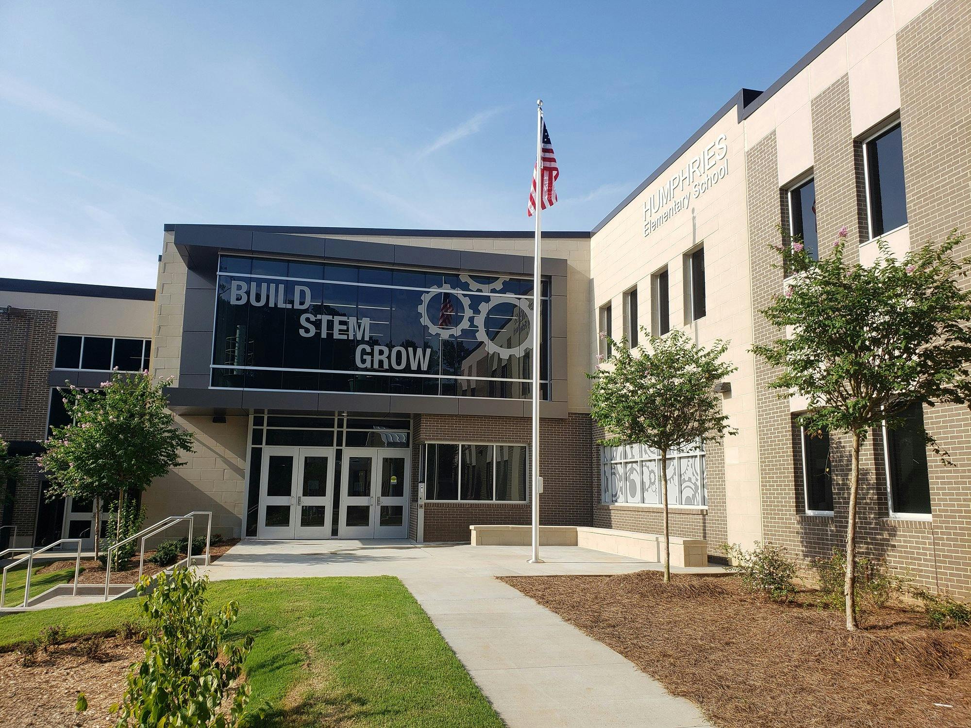 Front door of school with a paved walkway, flagpole, grass and trees. Entrance to the school shows a large glass mural with cog wheels and the words "Build, Stem, and Grow" below glass is two double doors.