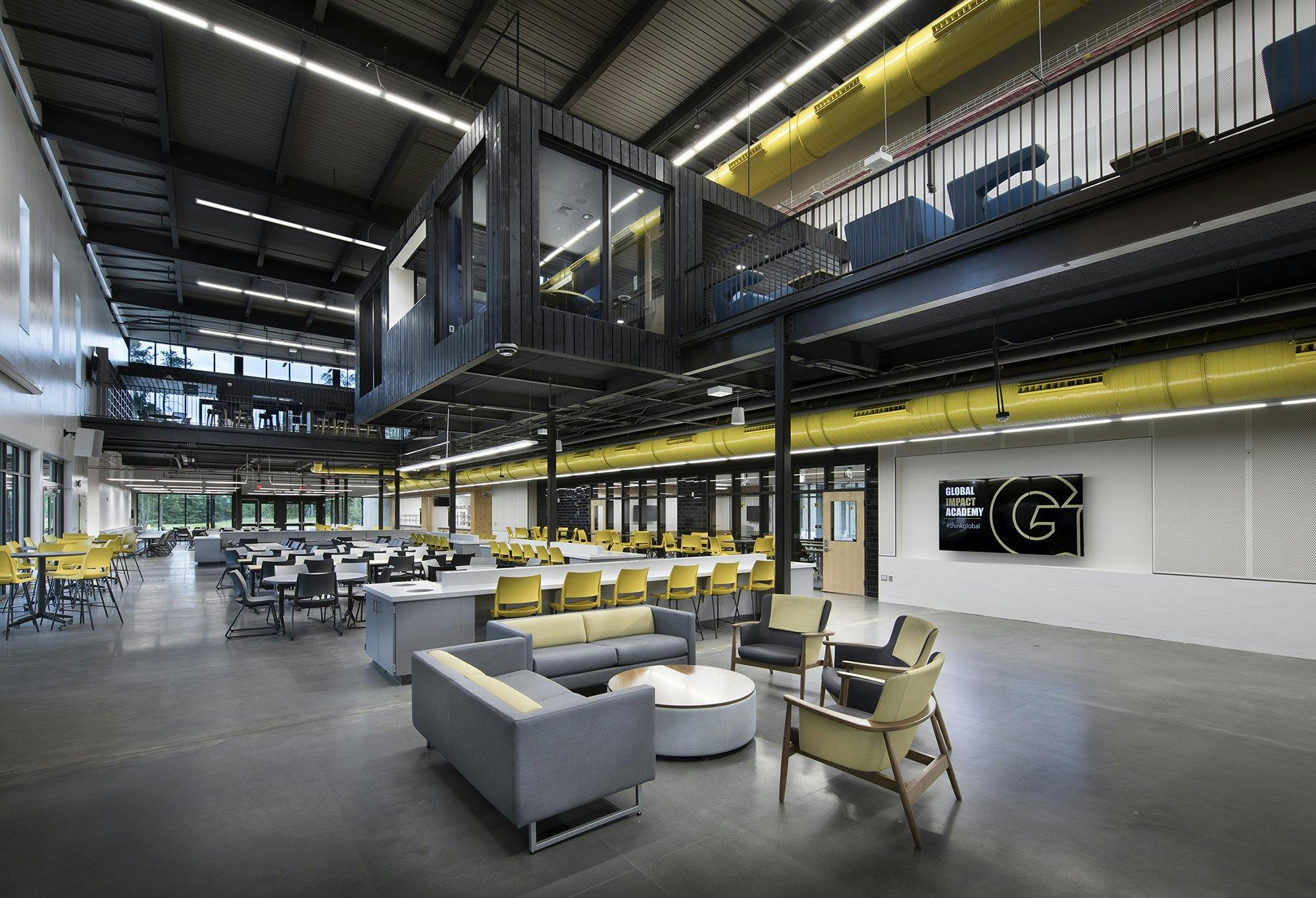 A dark wooden room overhangs a large open seating area filled with gray couches and yellow chairs. Dark exposed ceilings and painted yellow duct work are seen throughout.