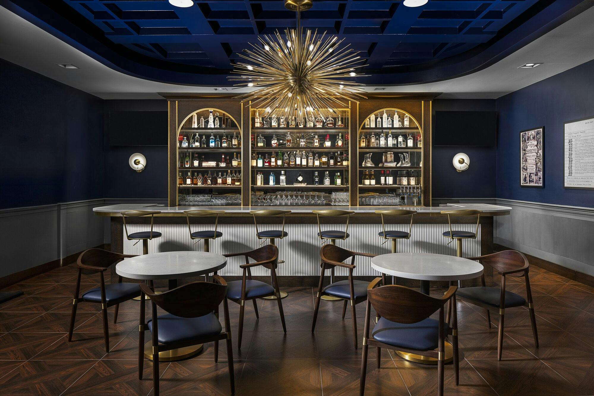 Intimate bar area with a navy blue ceiling and blue and white walls. A starburst chandalier above the bar with bar stools, tables and chairs.