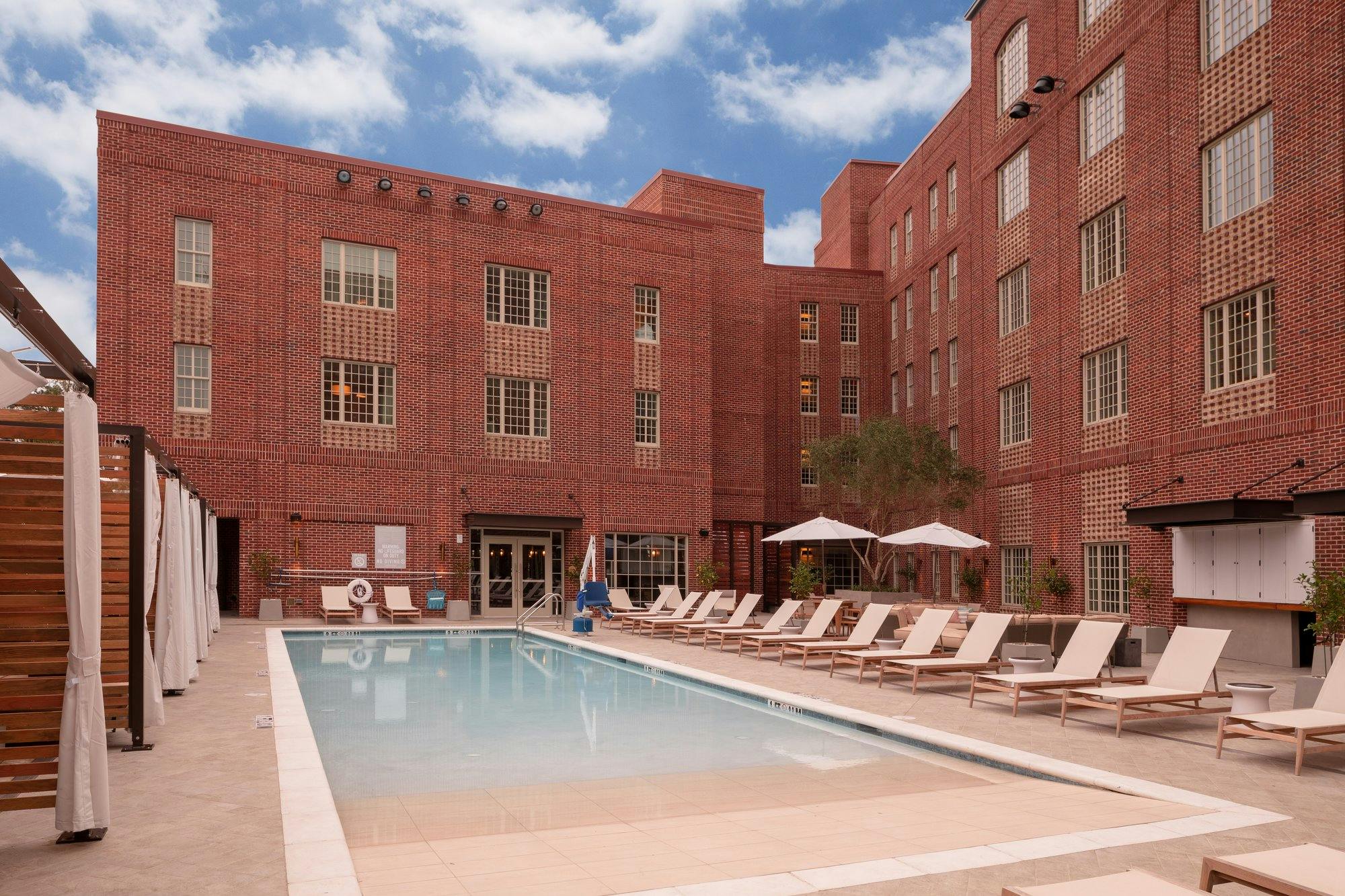 Hotel pool lined with white lounge chairs and umbrellas. Curtained cabanas and the brick exterior of the Alida Hotel surround the pool deck.