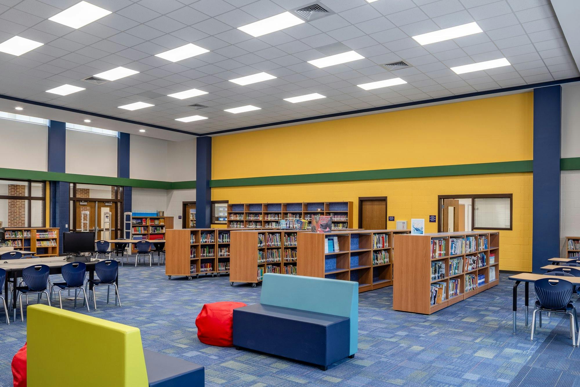 Short wooden shelves with many books line the aisles of an elementary school library. Cozy couches and chairs sit on a blue carpeted floor.