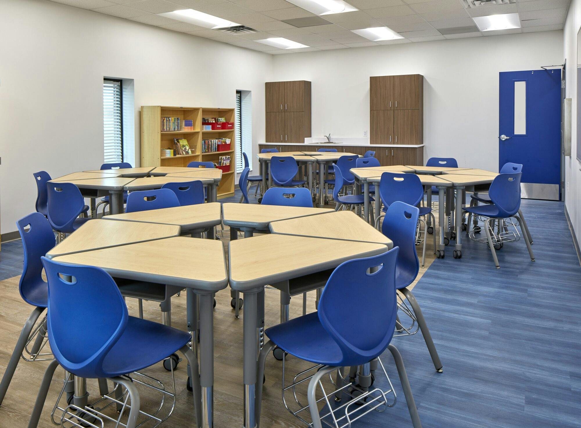 A classroom with blue and tan linolium, four sets of six desks forming circles with blue chairs and a blue door leading to a hall in the background.