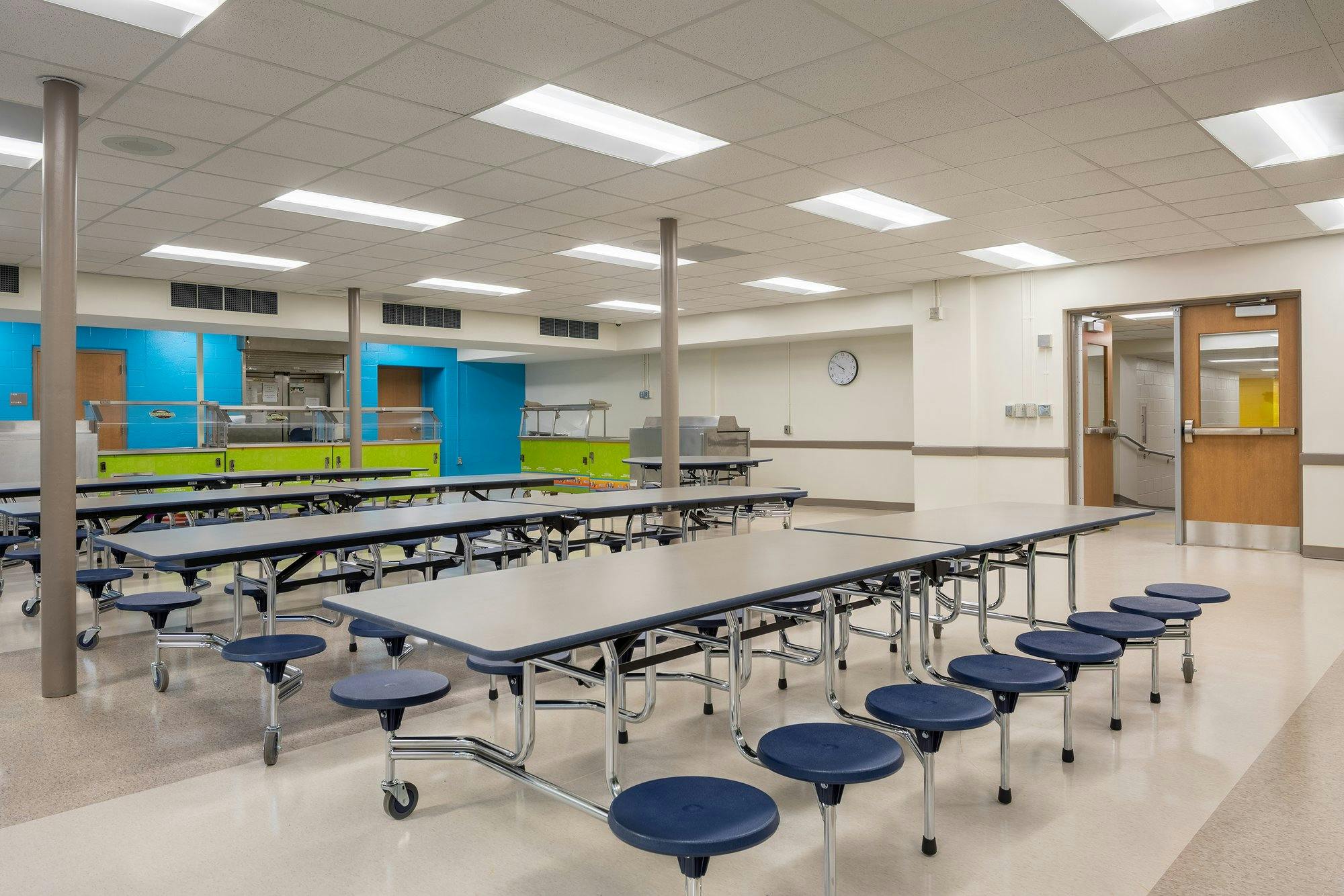 Bright cafeteria with electric blue and green accents. Many pedestal seats line groupings of tables.