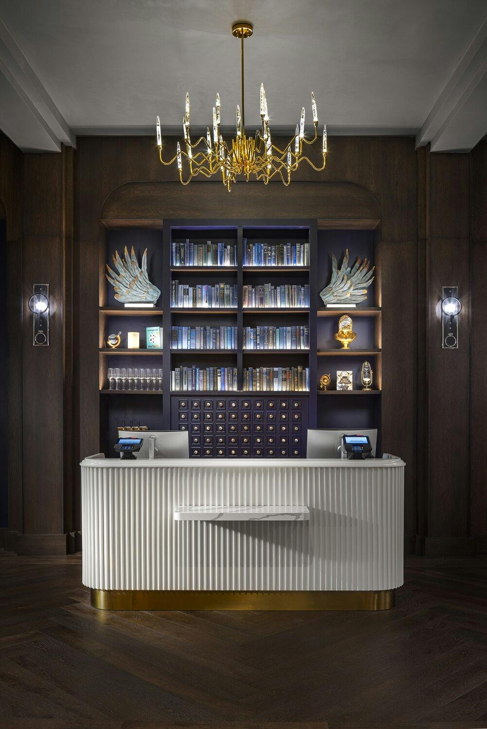 Guest services desk with white standing counter in front of a bookcase filled with books and side light sconces on either side.