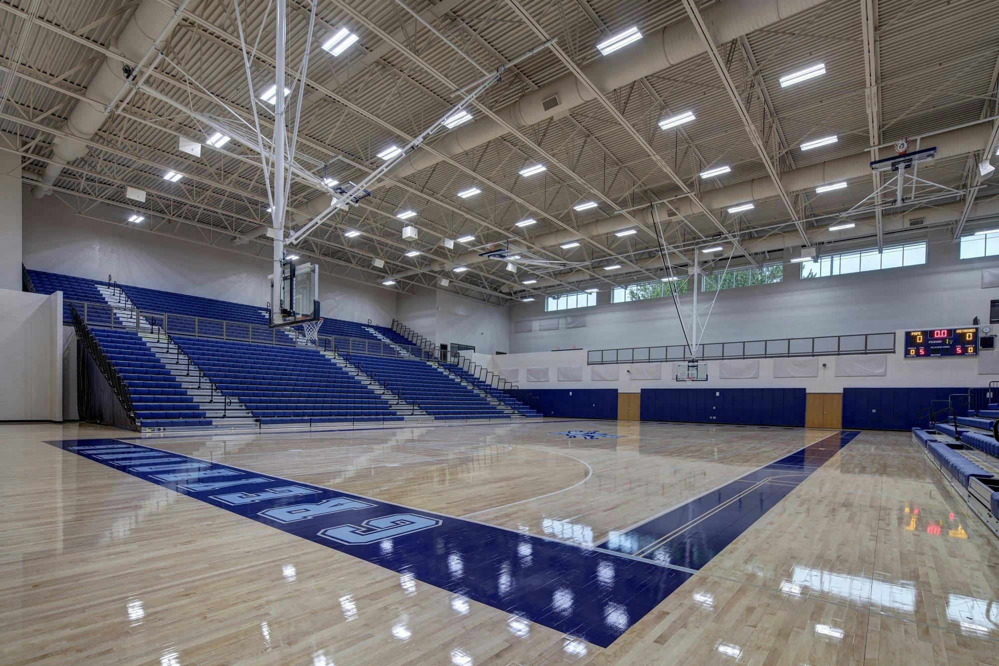 Blue bleachers surround a bright wood surfaced basketball court with blue painted accents.