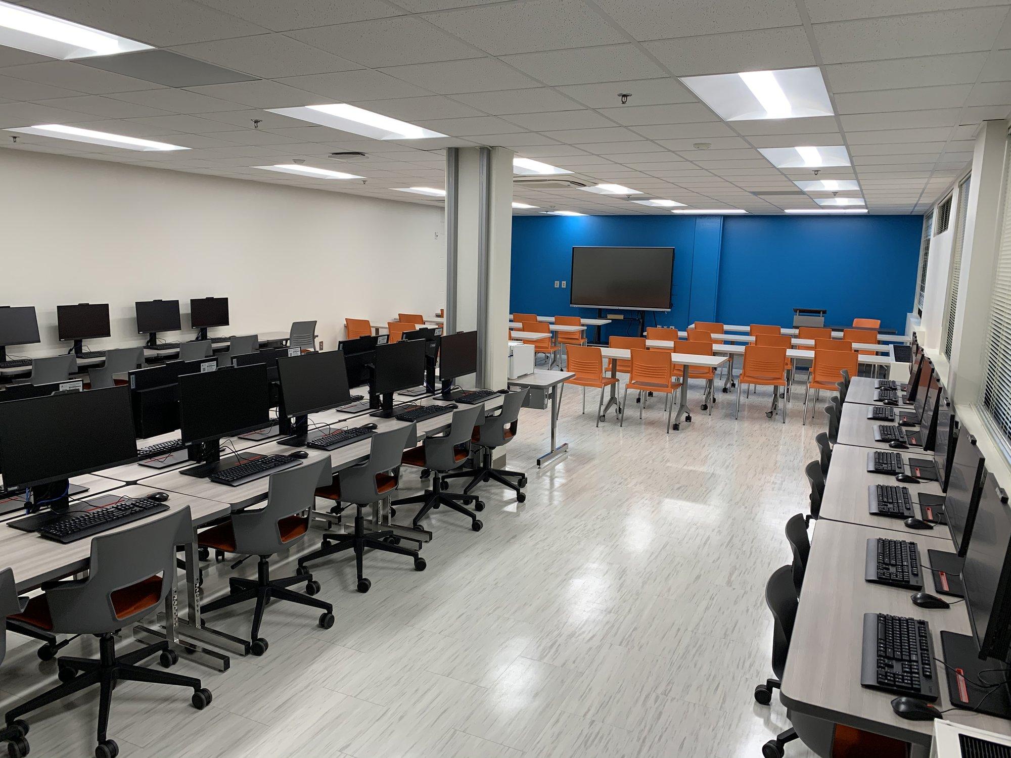 Office chairs are neatly lined up to many computer workstations. A presentation area with more seating can be seen in the back of the room.