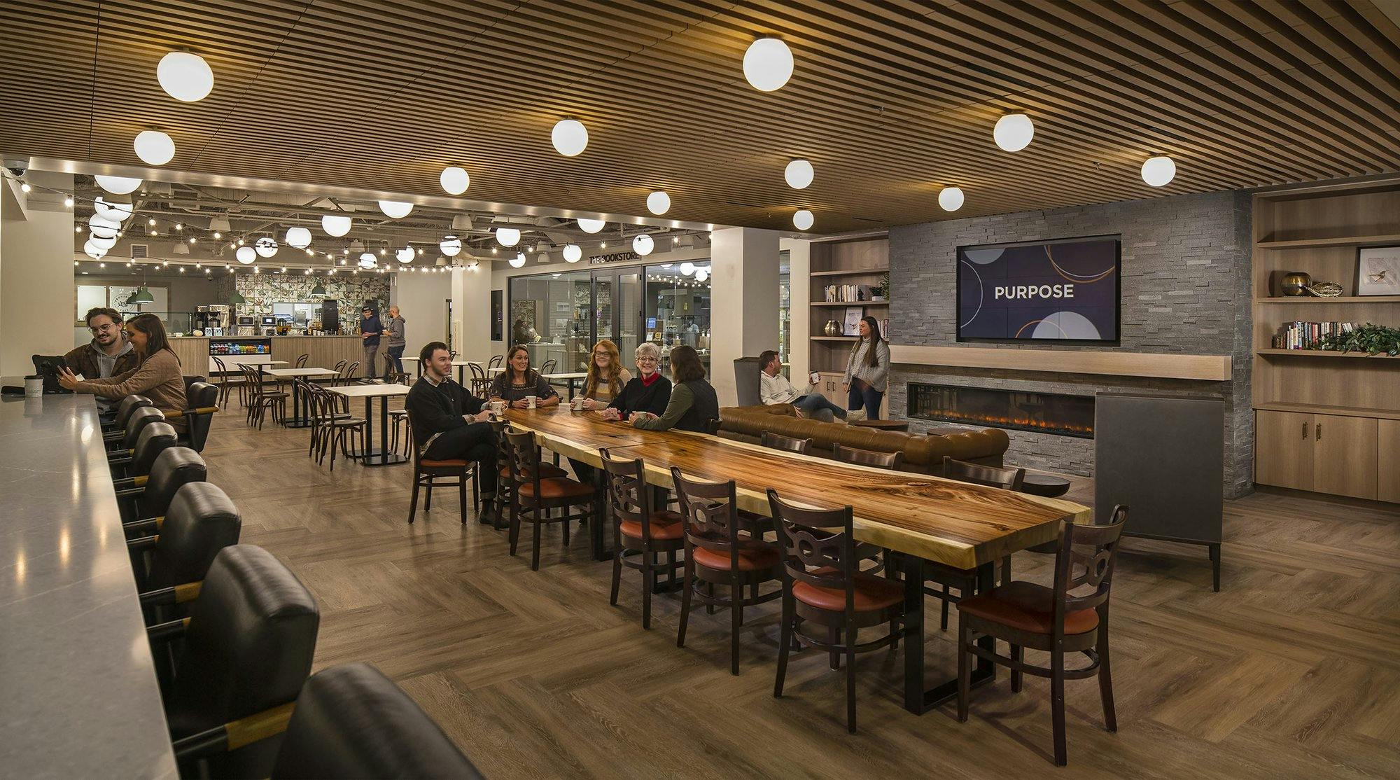 cafeteria seating area, featuring spherical light fixtures, a long wooden dining table, a fireplace, and a cafe in the background