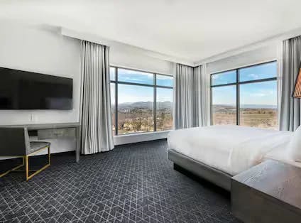 A spacious guest suite with dark colored textured carpet, neutral bed lines and cutains. Desk with chair and hanging tv. Suite has two windows at the corner of the room to maximize the views.