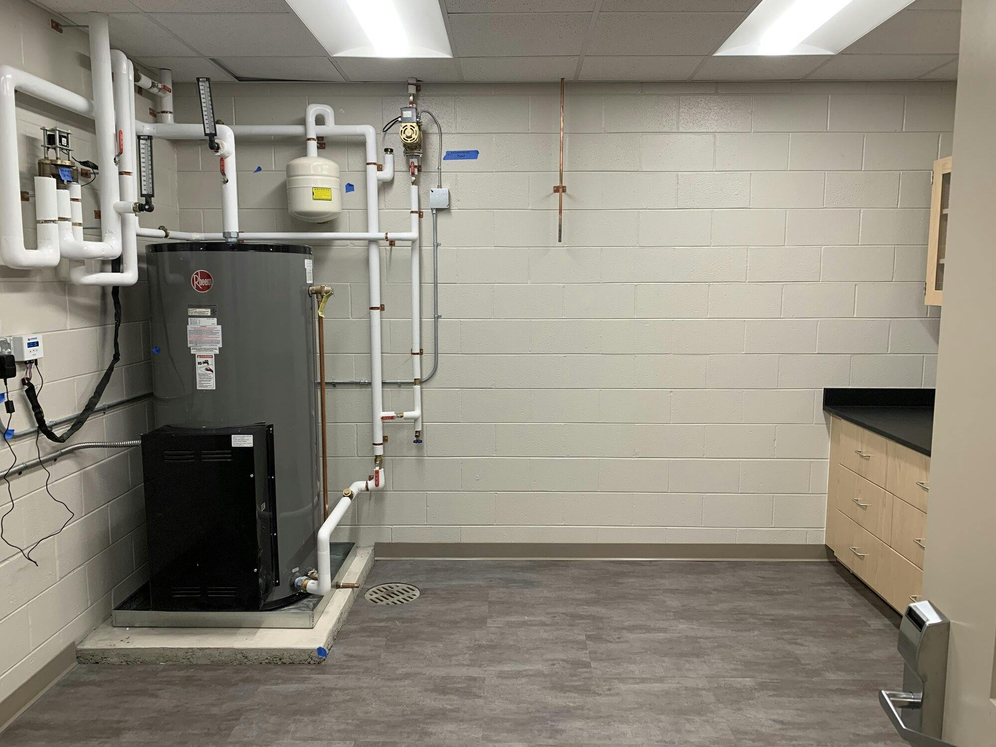 Room with Waterheater tank and plumbing lines feeding to the work station in adjacent room. Room also includes overhead cabinets, countertops and drawers for storage.