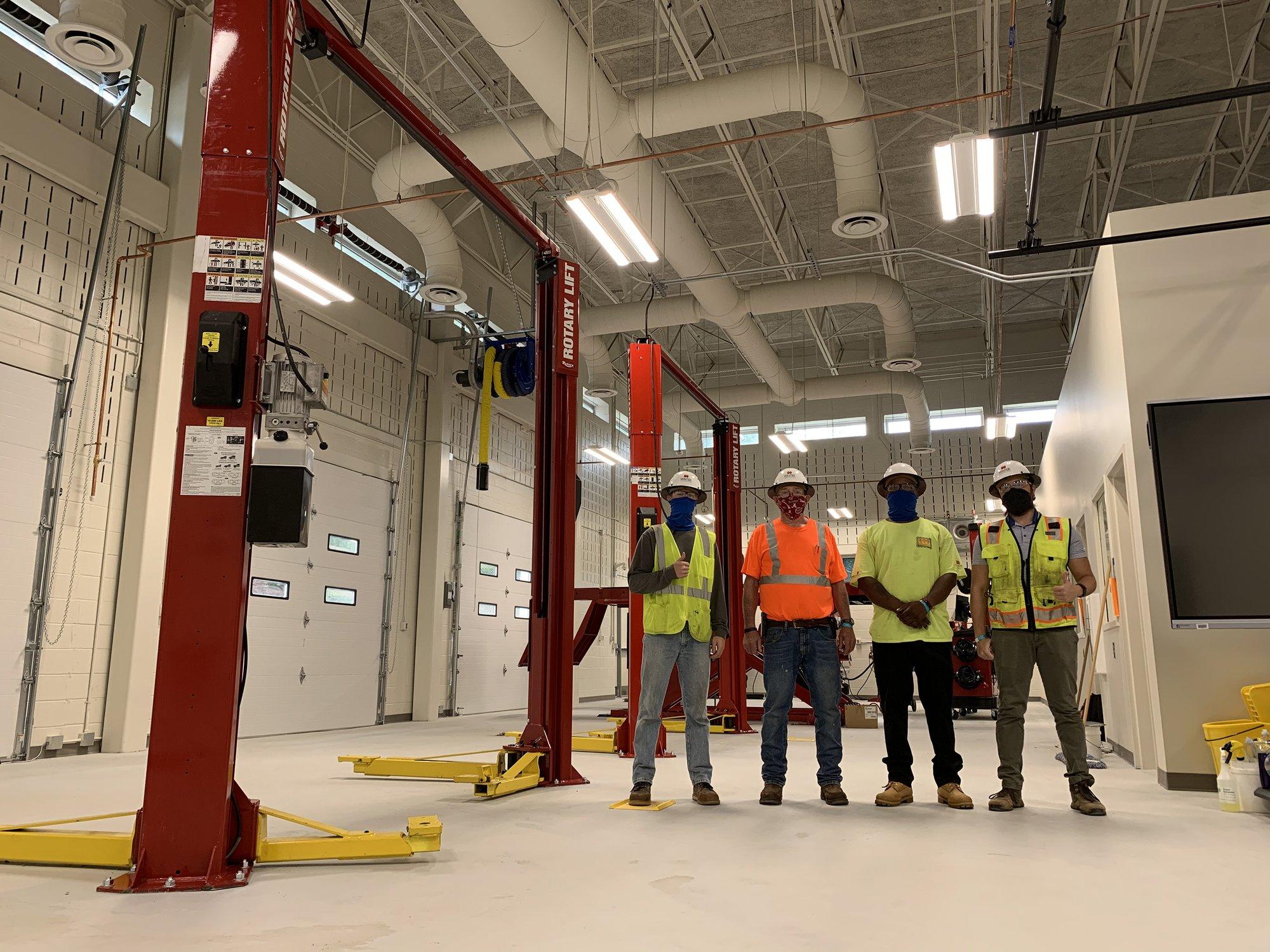 Four men in construction clothing stand in a garage with large red painted metal lifts.