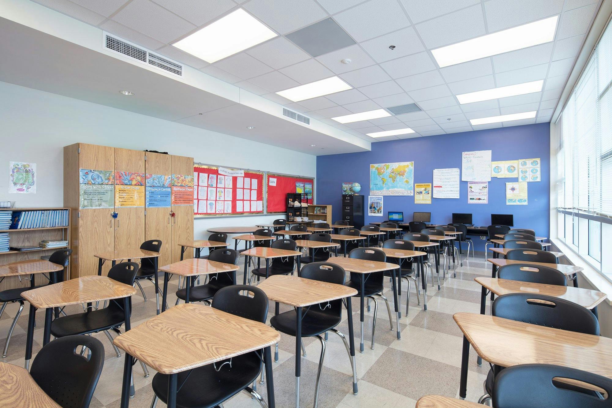 Desks and chairs fill a long classroom with a blue accent wall at the rear.