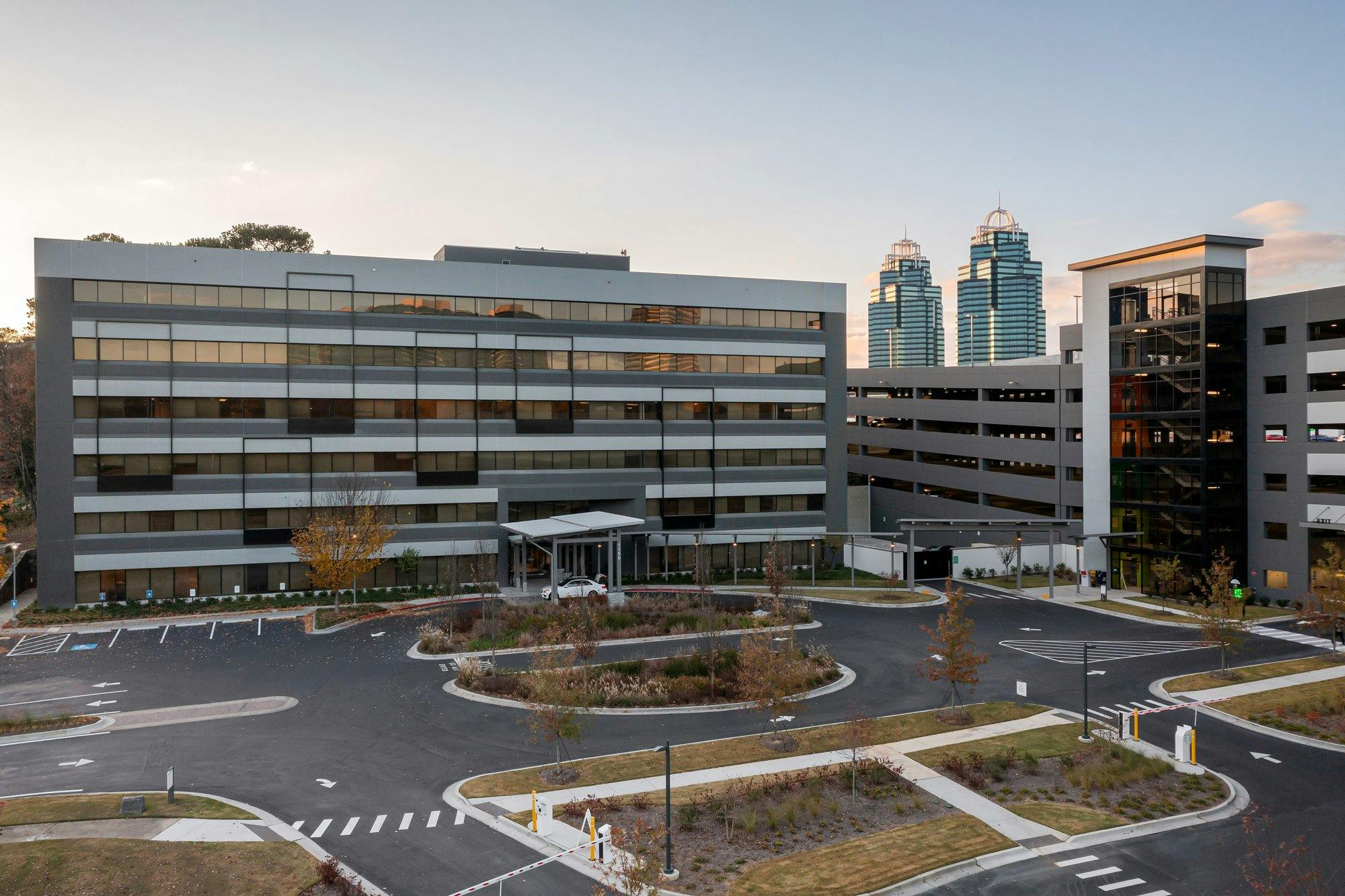 Building facade featuring lots of windows, a labeled parking lot and landscaping. In the background there is a clear image of the king and queen towers.