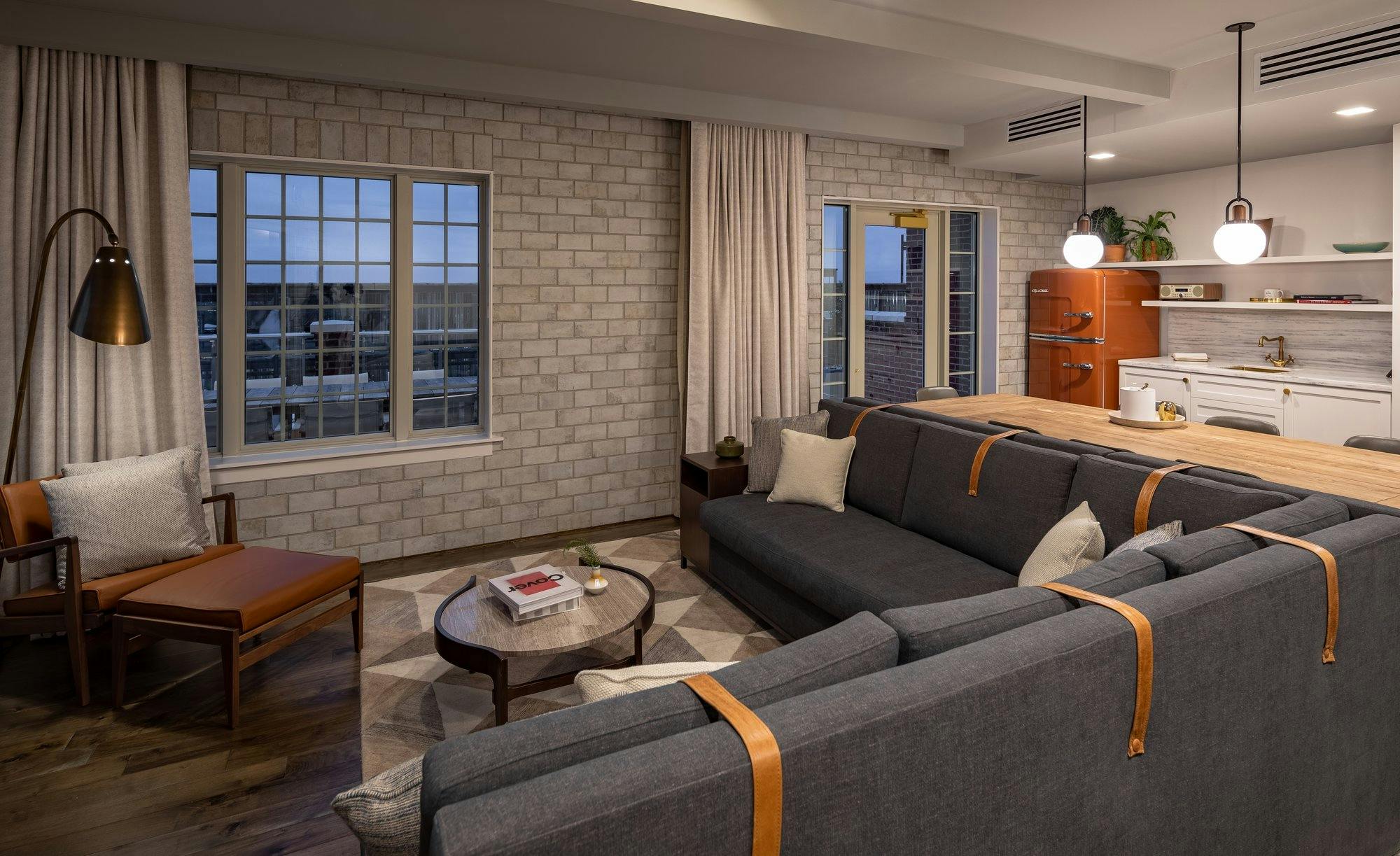 Living space of a hotel room at The Alida Hotel with a gray masonry wall, dark gray couch, and leather accents. A kitchenette with a rust colored retro refrigerator can be seen in the back of the room.
