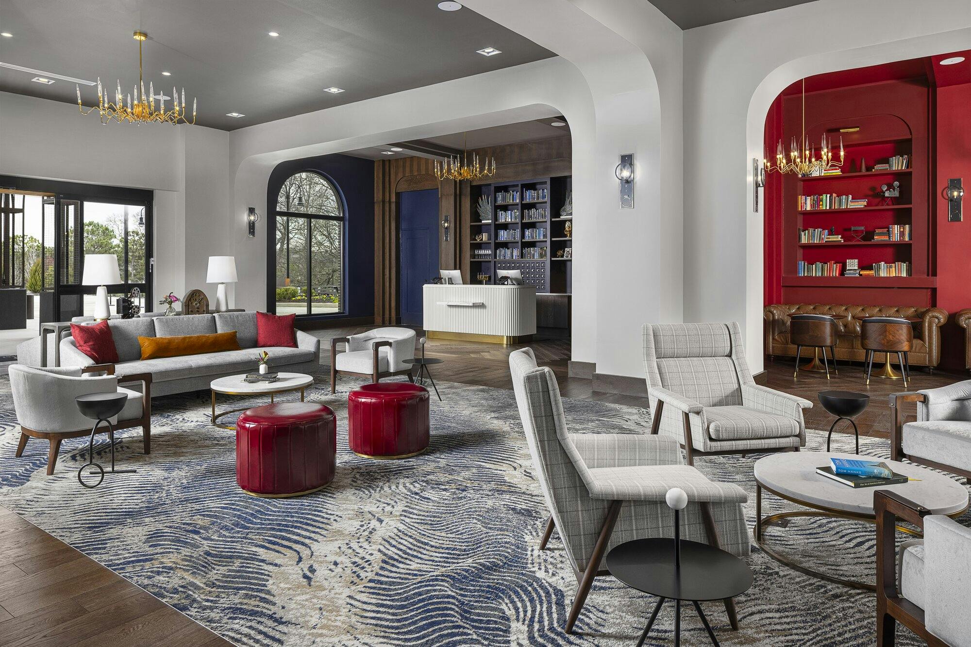 Waiting lobby with curved archways and chandaliers. Multiple seating areas; neutral colored couch and chairs with red ottomans. Opening up to guest services desk.