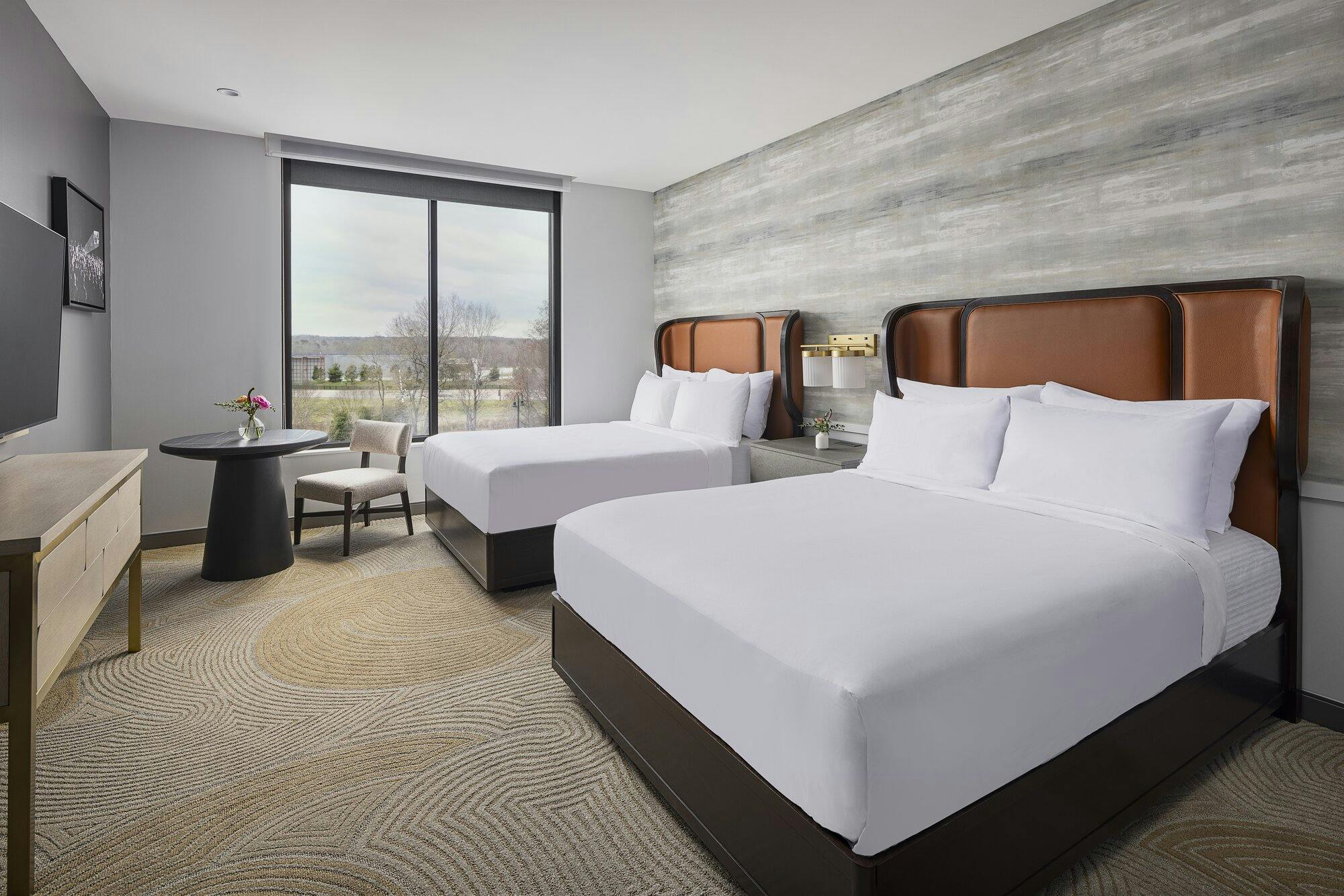 Guest suite with two queen beds with curved leatherbound headboards, white linens and pillowcases. Patterned textured neutral tone carpet with a table and chair, dresser and hanging TV. A large window on one wall overlooking trees.