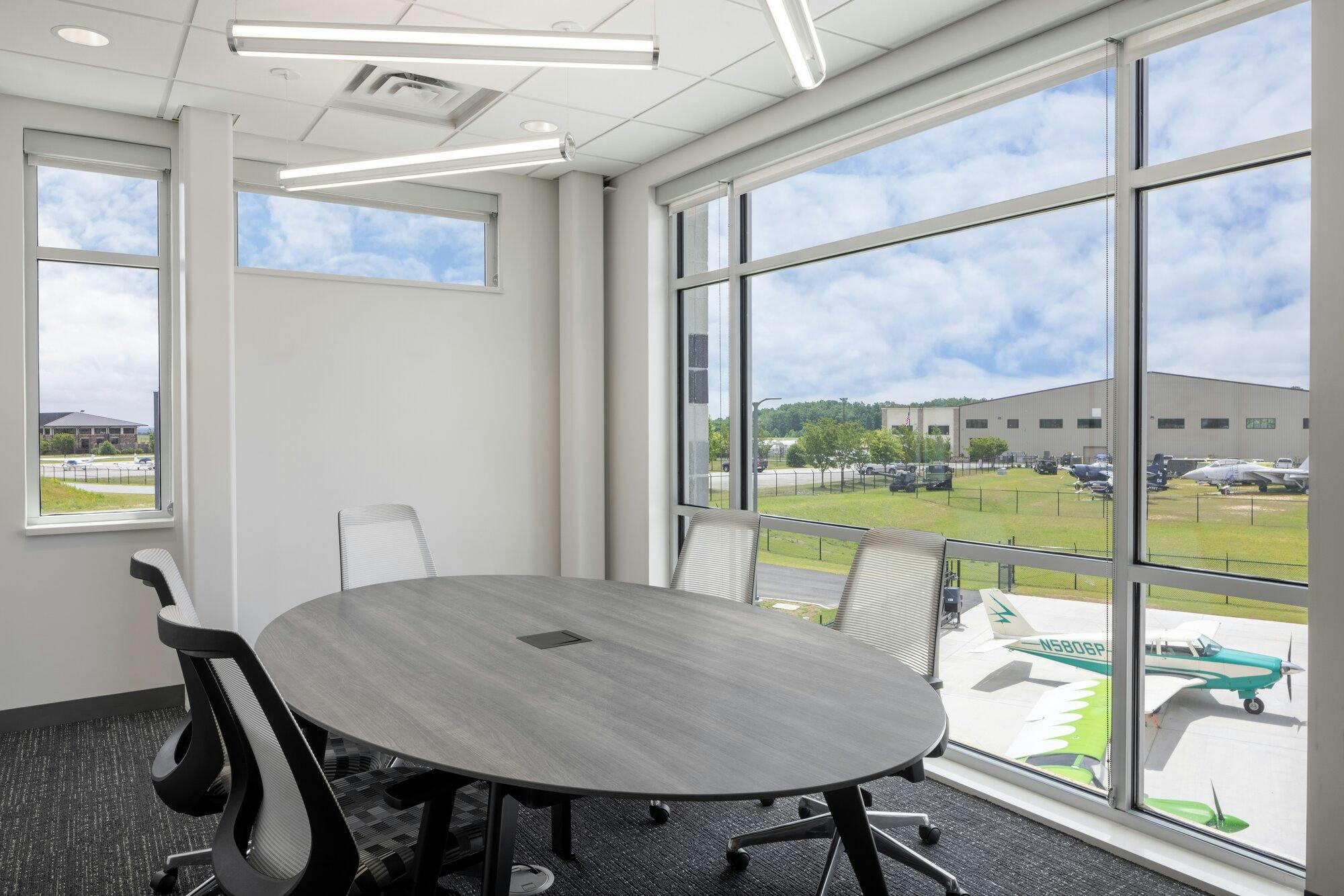 Classroom image with one wall as a window overlooking the airplane hangar and airplanes parked. The room has a round table surrounded by chairs and light bars hanging from the ceiling.