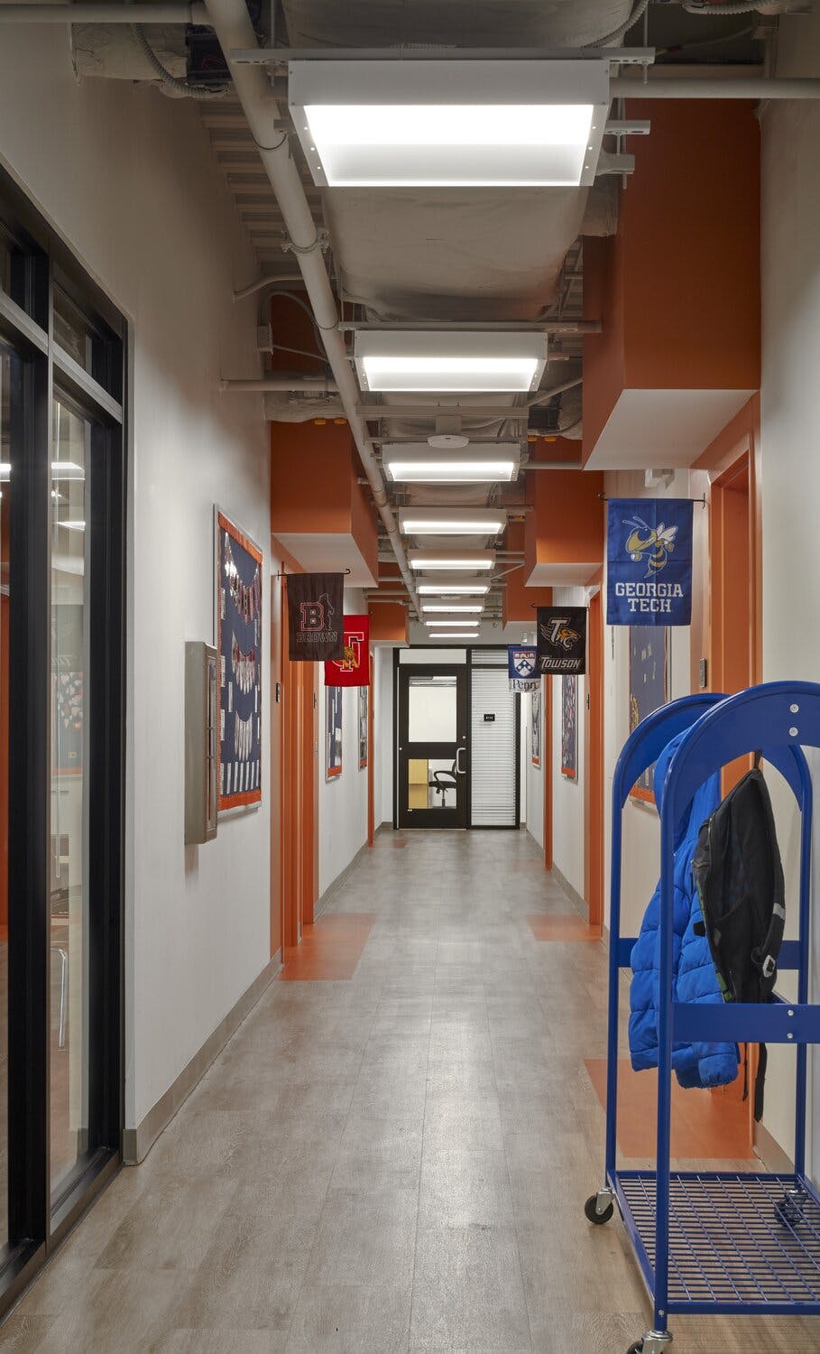 A hallway with linolium floors, orange an white painted walls, and square overhead light boxes leading to classrooms.