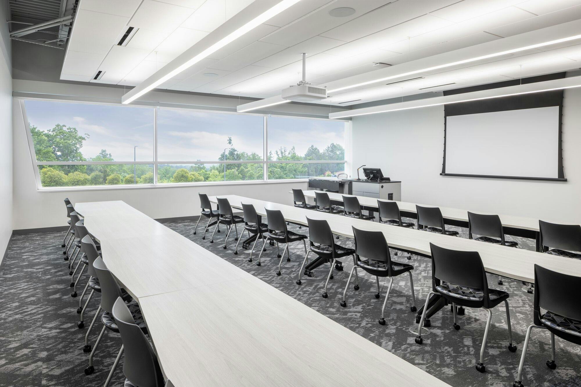 A classroom with one wall of windows, long tables with chairs. A projector hanging from ceiling and light bars that stretch the width of the room above each table.