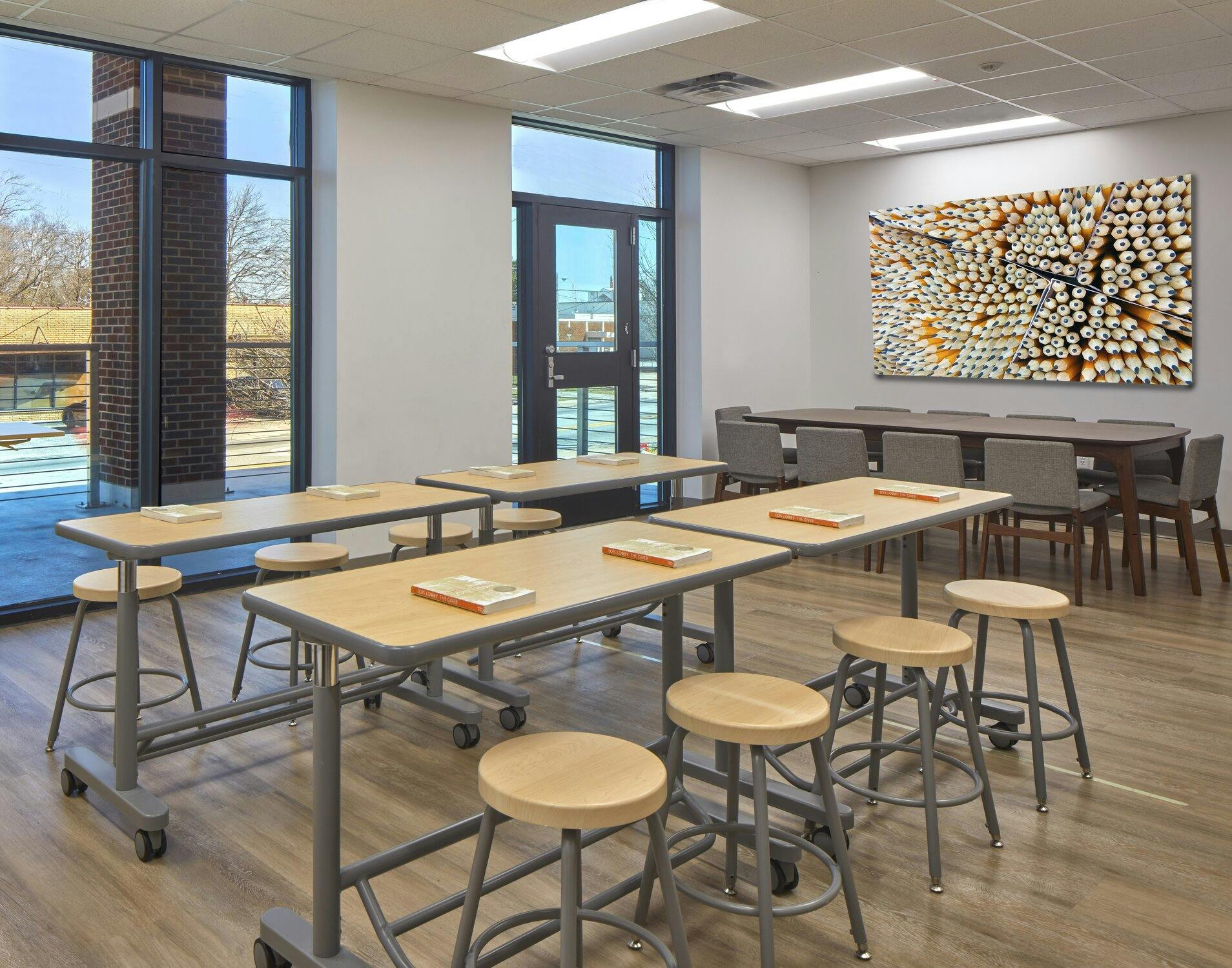 A classroom with standing desks and stools under them. A long table with chairs in the background along with two floor to ceiling windows and a door leading outside a few feet away.