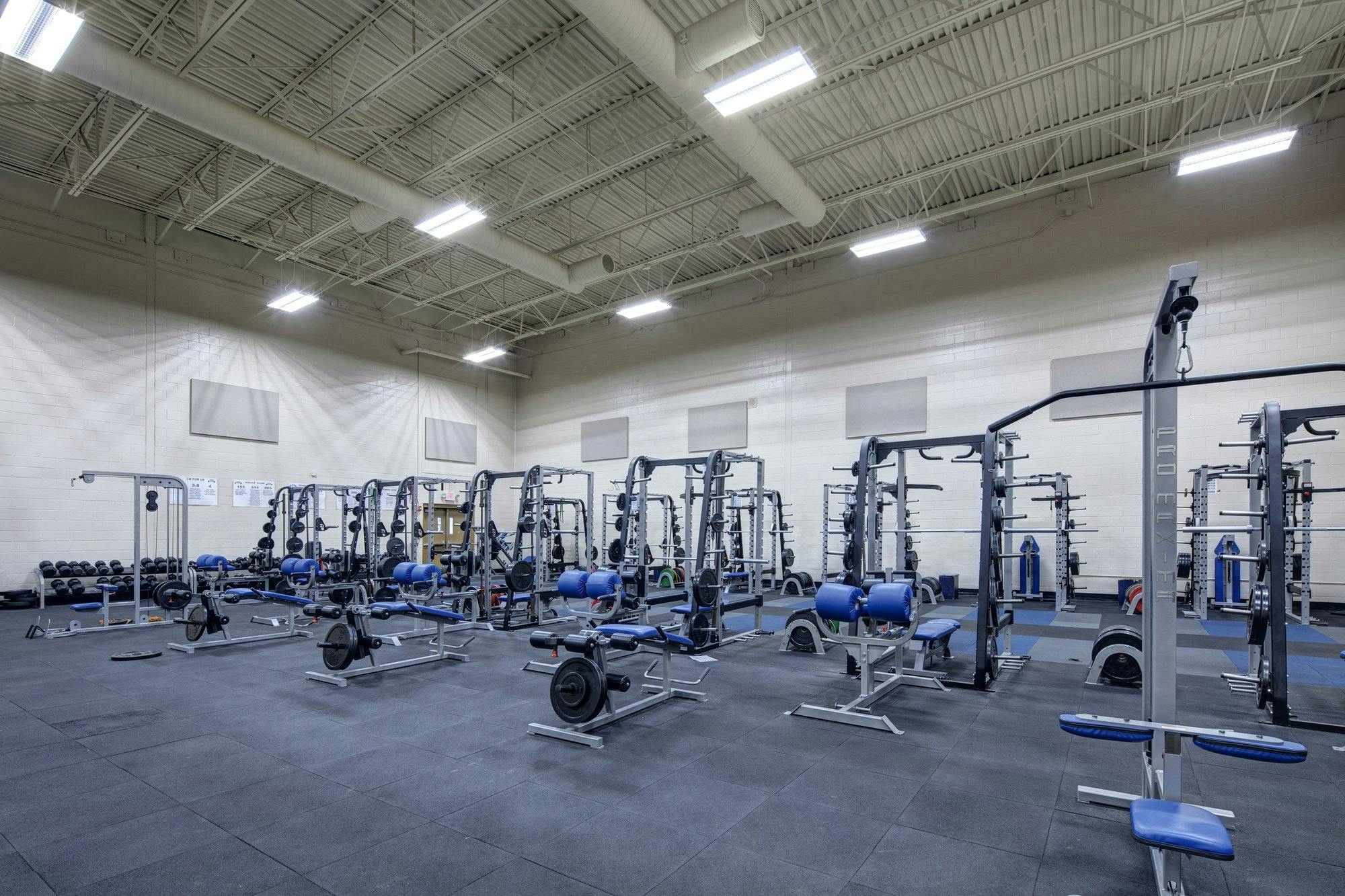 Different types of weigh lifting and exercise equipment dot the floor of a large room with high white exposed ceilings.