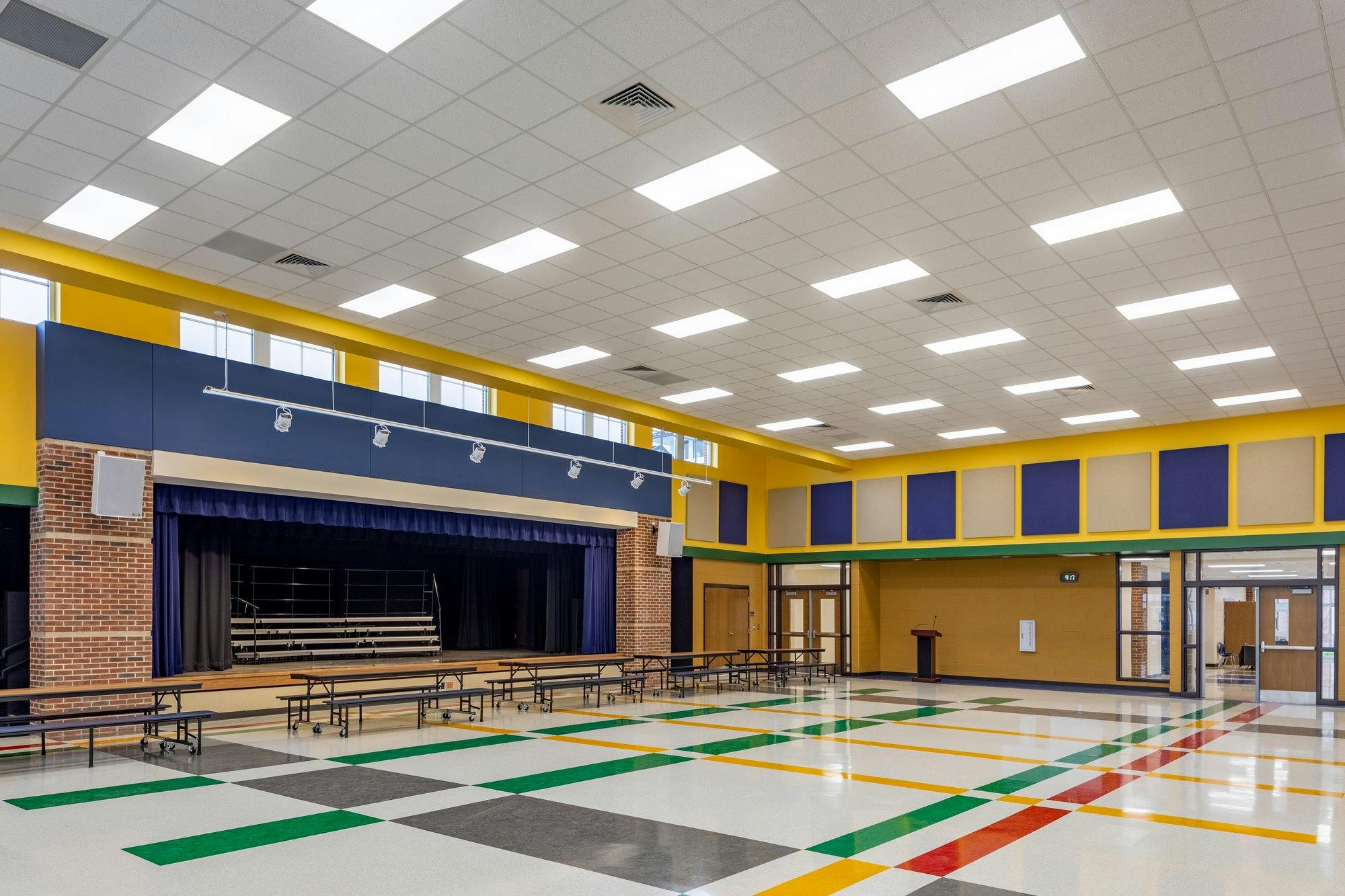 A multi-color tile floor dots a large open cafeteria/auditorium with a curtained stage off to the side.