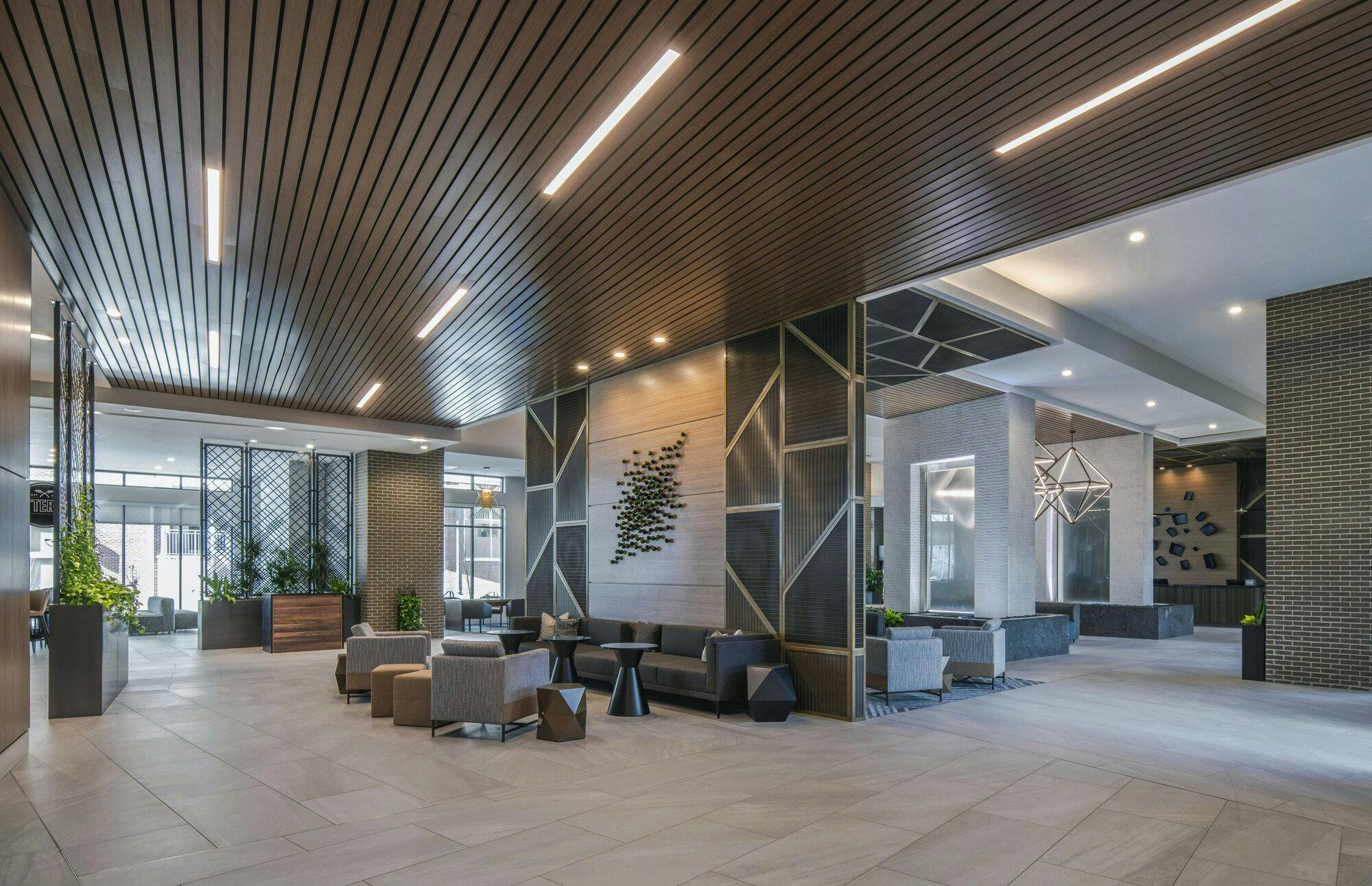 Large lobby area with wood panels on the ceiling mixed with light bars and recessed lights. Multiple seating arrangements in various locations.