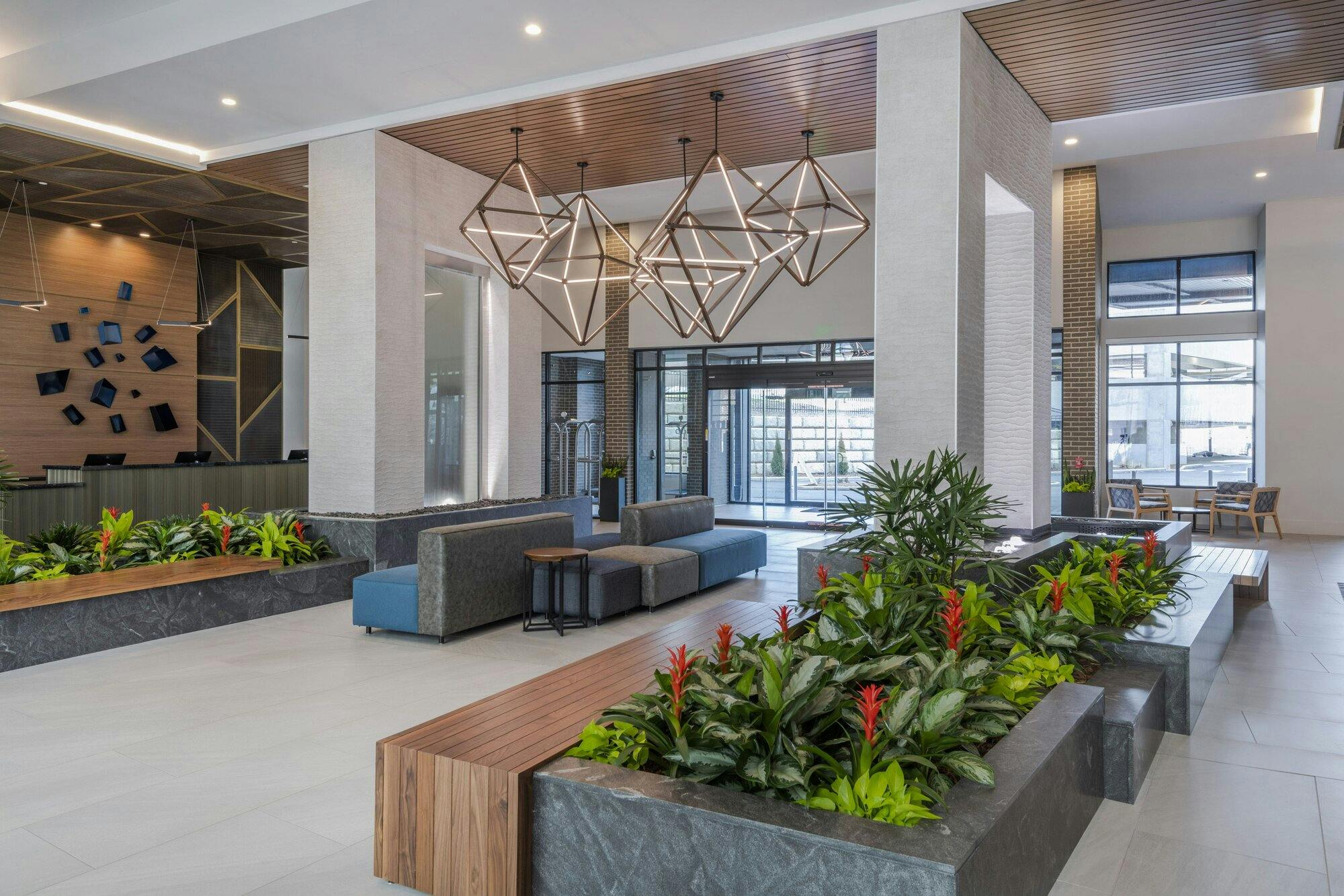 Foyer area, next to guest services showing four diamond shaped chandaliers at different sizes and heights grouped together. Walls with built in waterfalls stand on both sides of chandaliers surrounded by wood seating, lights, and foilage.