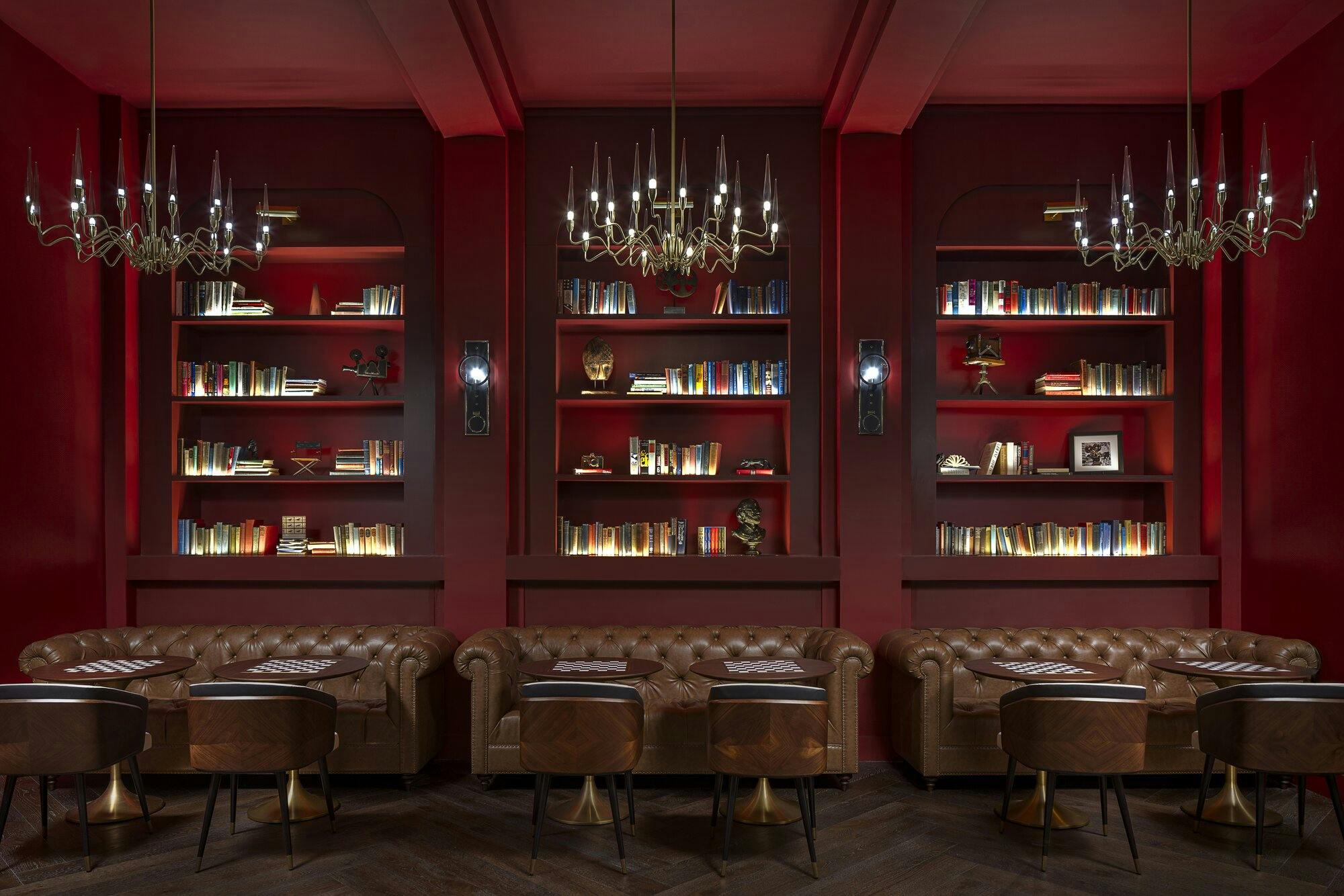 Lounge area with deep red walls and ceiling with three chandaliers. Built in bookcases filled with books and three large cognac colored leather couches with tables and leather chairs.