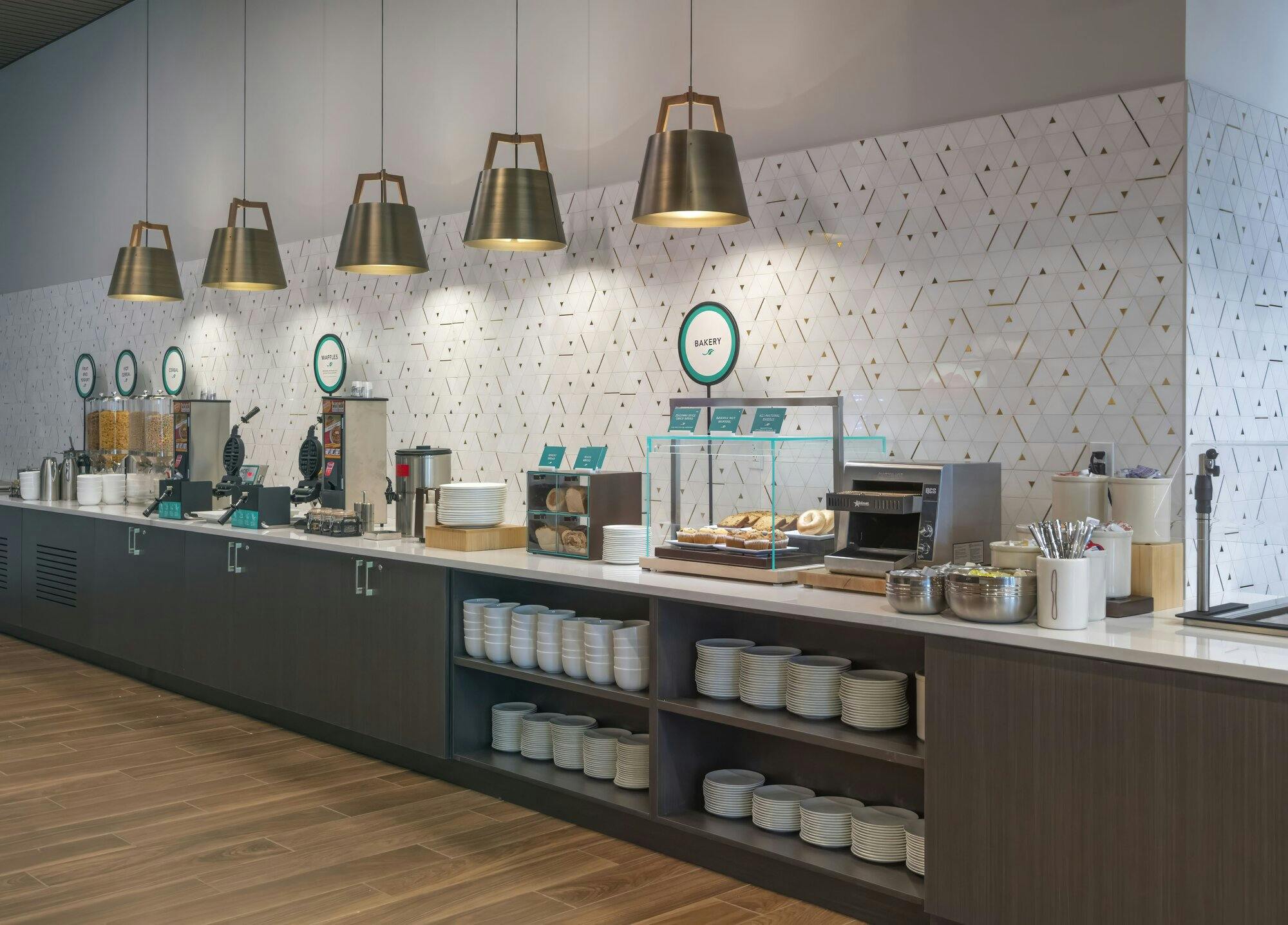 Breakfast area with hardwood floors, a geometric backsplash and brass hanging pendant lights shining down on a breakfast bar. Bar has a white counter top with multiple displays to hold pastries, kitchen appliances, cutlery, and cereal option self serve containers. Shelves and cabinets underneath.