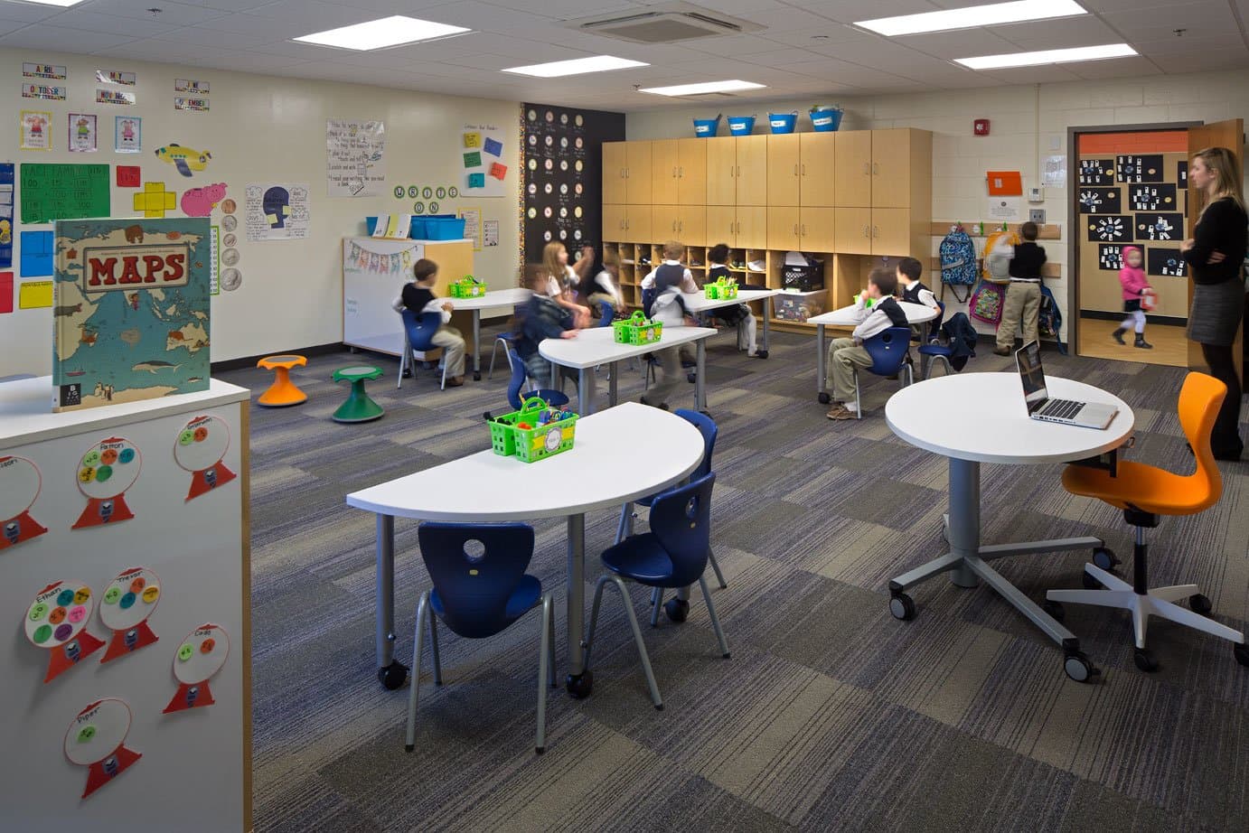 Children seen sitting in a classroom with white tables and wooden cabinetry.