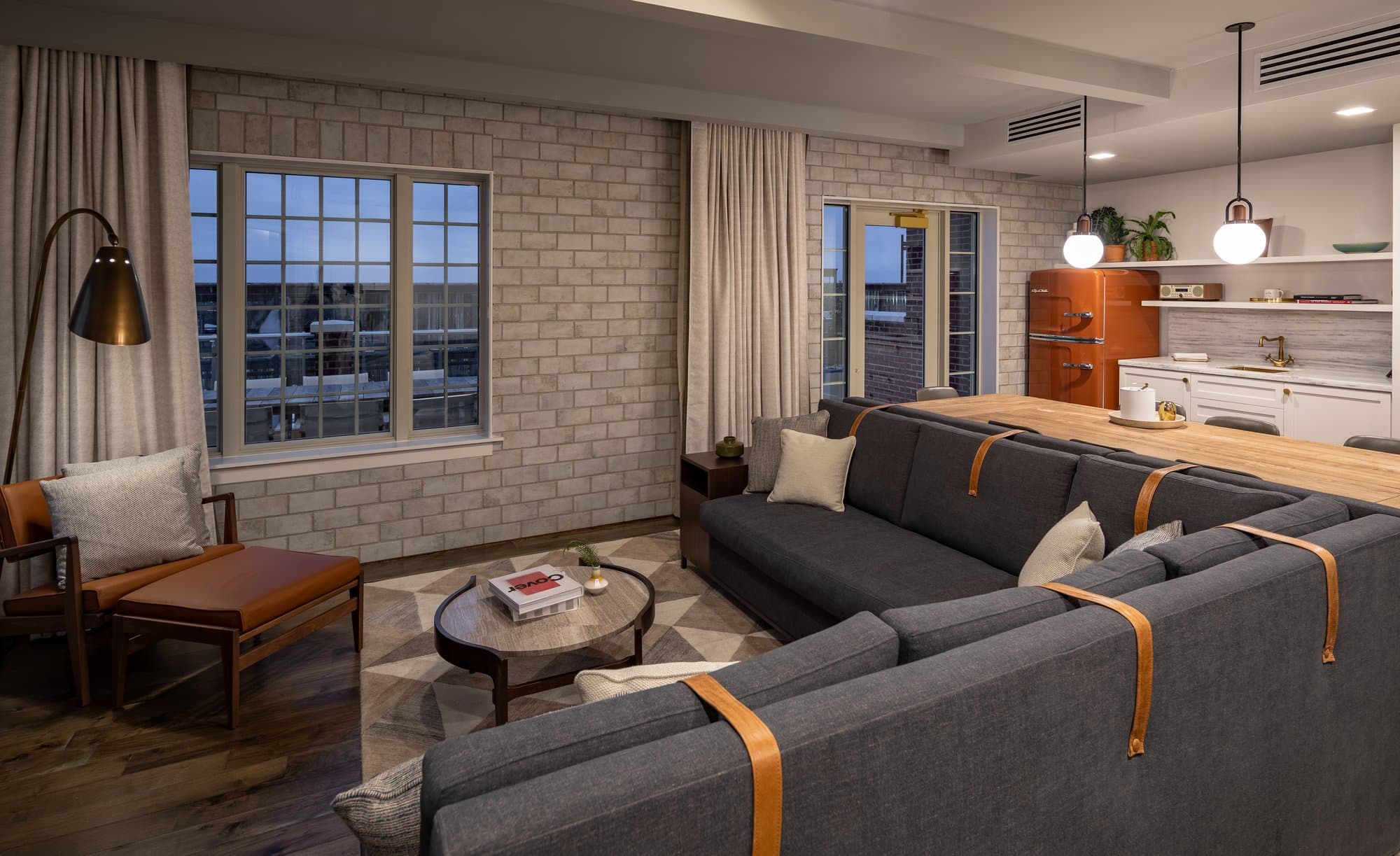Living space of a hotel room at The Alida Hotel with a gray masonry wall, dark gray couch, and leather accents. A kitchenette with a rust colored retro refrigerator can be seen in the back of the room.