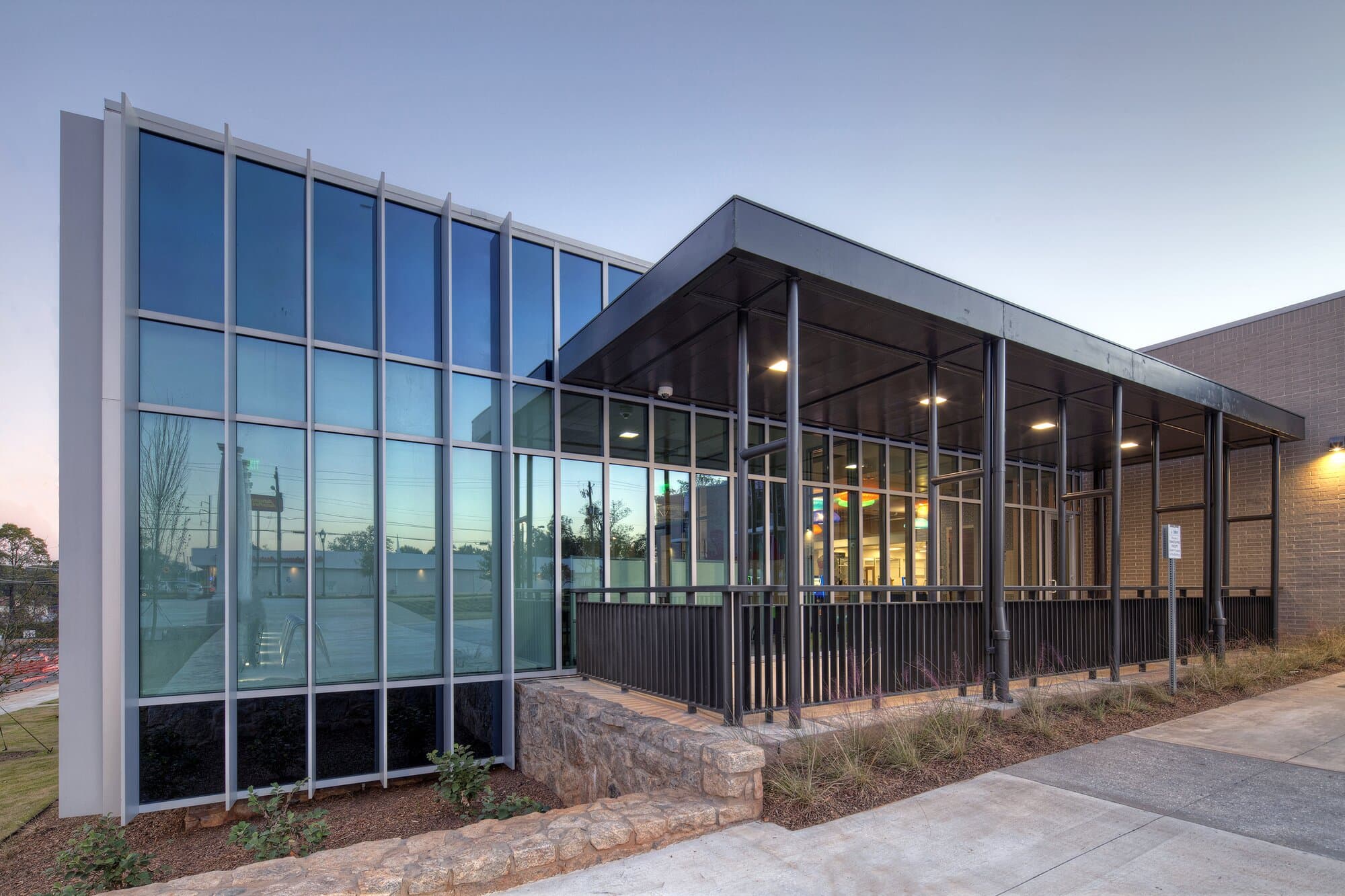 Entrance to facility with large glass windows, lighting, paved entrance walkway, and metal awning.