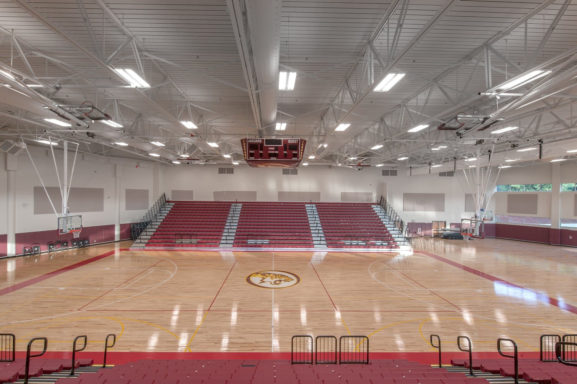 Rows of red bleachers surround a large gymnasium basketball court with light wooden floors and painted red lines.