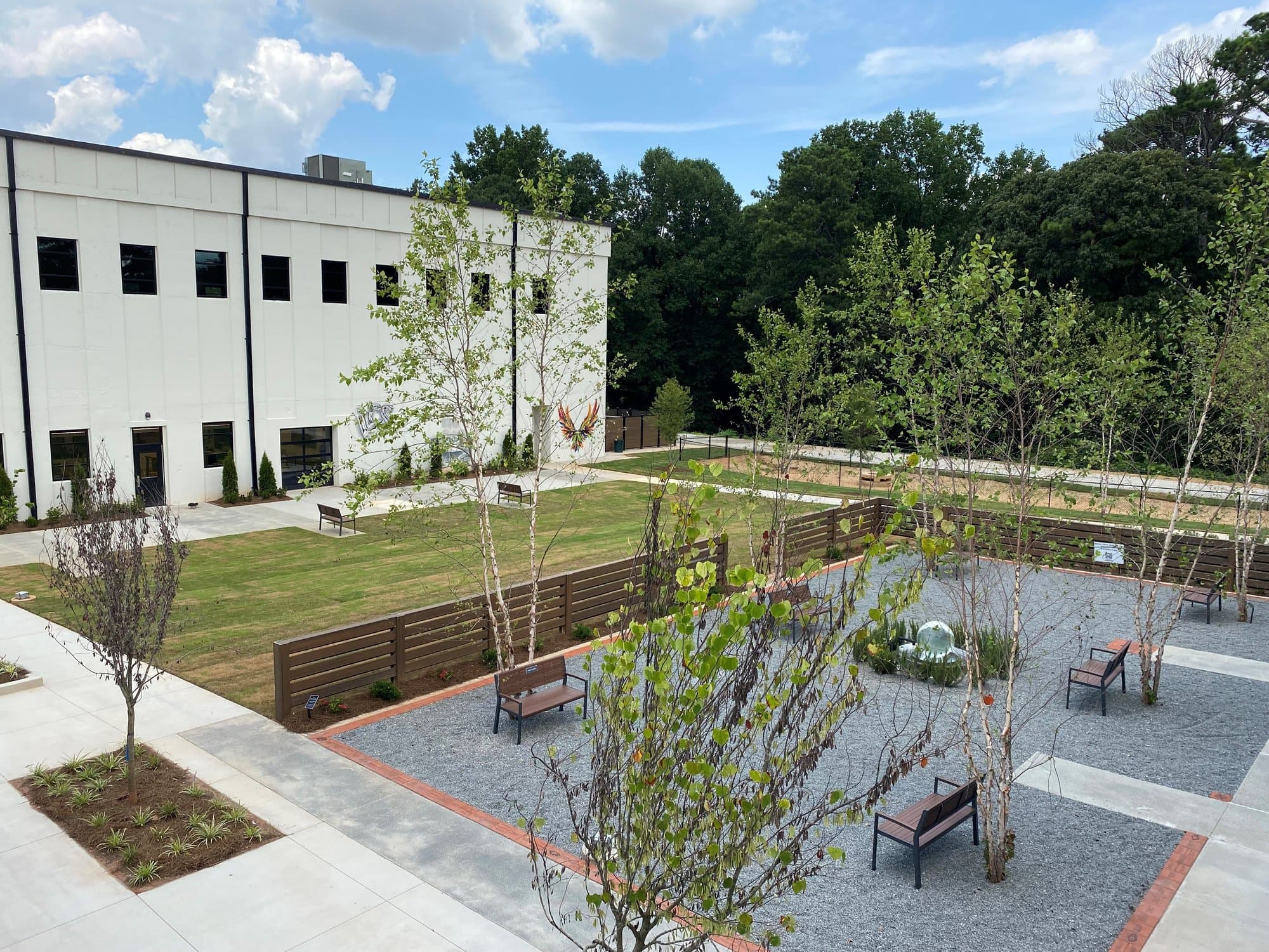 A tree-lined gravel courtyard with benches in front of a white-exterior two-story building.